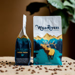 12 oz. Colombian coffee bag with a mountain scene and illustration of a Elk with Wild Rivers Logo. Tasting notes for this coffee include: Caramel, Floral, Red Apple. Front and side view of bag on coffee table
