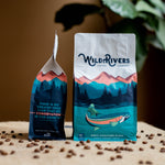 12 oz. Wild Rivers Signature Blend coffee bag with a mountain scene and illustration of a rainbow trout with Wild Rivers Logo. Tasting notes for this coffee include: Smooth, Chocolate, Berries. Front and Side view on coffee table