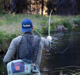 Man fishing facing away from the camera and towards the river. He is wearing a Navy Blue Snap Back hat on backwards so the viewer can see the logo. The man is casting his line into the water.